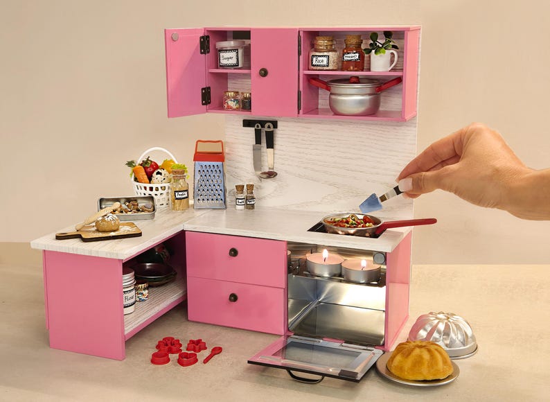 K&ouml;nnte beinhalten: Eine Miniatur-K&uuml;chenzeile in Rosa mit offenen Schr&auml;nken, einem Herd und einem Backofen. Das Set enth&auml;lt einen Korb mit Gem&uuml;se, Gew&uuml;rzgl&auml;ser und einen kleinen Topf. Eine Hand r&uuml;hrt mit einem Spatel Essen in einer Pfanne auf dem Herd um.