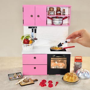K&ouml;nnte beinhalten: Eine Miniatur-K&uuml;chenzeile in Rosa mit Herd, Schr&auml;nken und Zubeh&ouml;r. Das Set enth&auml;lt eine Bratpfanne, einen Korb mit Gem&uuml;se und verschiedene Backutensilien. Eine Hand h&auml;lt einen Pfannenwender &uuml;ber der Pfanne.