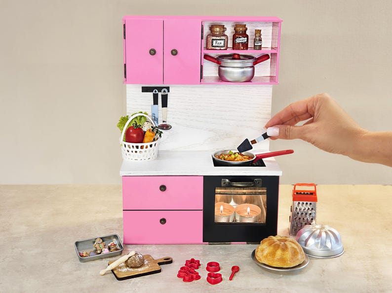 K&ouml;nnte beinhalten: Eine Miniatur-K&uuml;chenzeile in Rosa mit Herd, Schr&auml;nken und Zubeh&ouml;r. Das Set enth&auml;lt eine Bratpfanne, einen Korb mit Gem&uuml;se und verschiedene Backutensilien. Eine Hand h&auml;lt einen Pfannenwender &uuml;ber der Pfanne.