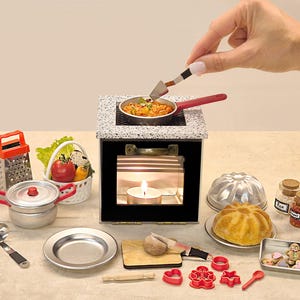 Puede incluir: Escena de cocina en miniatura con un pequeño horno, una sartén con pasta y varios utensilios de cocina. Incluye un rallador, una olla, un plato y una cesta de verduras. Una vela arde dentro del horno. También hay moldes para hornear y especias.
