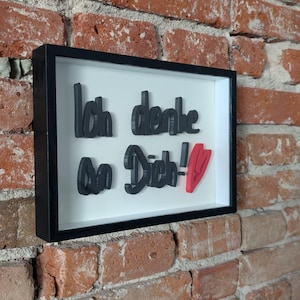 Könnte beinhalten: Gerahmtes Wandkunstwerk mit dem deutschen Satz "Ich denke an Dich!" in schwarzer 3D-Schrift, mit rotem Herzakzent. Das Kunstwerk ist vor einem weißen Hintergrund platziert und schwarz gerahmt, an einer Backsteinmauer befestigt.