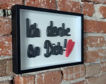 May include: A framed wall art piece with the German phrase "Ich denke an Dich!" in black 3D lettering, with a red heart accent. The artwork is set against a white background and framed in black, mounted on a brick wall.