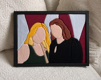 May include: A framed fabric wall art piece featuring two faceless figures. One figure has long yellow hair and wears a dark green top, while the other has brown hair and wears a black top. The background consists of vertical light blue and maroon stripes.