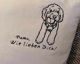 May include: White throw pillow with a black embroidered illustration of a dog holding a ball. The text "Mama, Wie lieben Dich!" is embroidered below the dog. The pillow is on a gray textured surface.