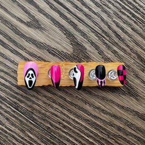 May include: A set of six press-on nails with a spooky theme. The nails are displayed on a wooden holder. Designs include a pink and black Ghostface mask, solid pink, a black and white Ghostface design, black with pink drips, and a pink and black checkerboard pattern.