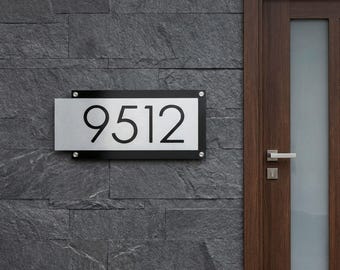 May include: A modern address sign with the number "9512" in black, set against a brushed metal background. The sign is framed in black and mounted on a dark gray stone wall, near a wooden door.