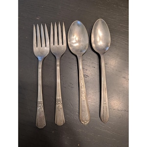 May include: Four pieces of vintage silverware, including two forks and two spoons, are arranged on a dark surface. The silverware is silver-toned, with decorative patterns on the handles. The forks have four tines, and the spoons have oval bowls.