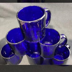 May include: A set of six cobalt blue glass mugs arranged in a stacked pyramid formation. Each mug features a smooth, cylindrical body with a sturdy handle attached to the side. The glass is translucent and deep blue, reflecting light on the surfaces.