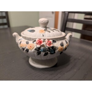 May include: A white ceramic trinket dish with a lid, adorned with hand-painted floral designs in pink, yellow, and blue. The dish has a pedestal base and small handles on either side. The lid features a small knob.