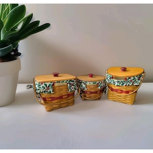 May include: Three woven baskets with light wood lids and red knobs. Each basket features a fabric liner with a green holly and red berry pattern, a red woven accent band, and a swing handle. The baskets vary in shape, including rectangular and round.