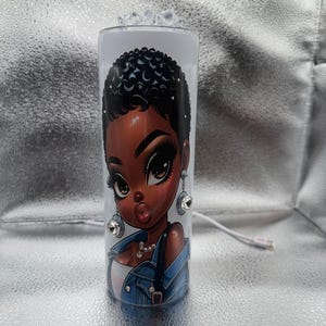 May include: A white tumbler featuring a cartoon illustration of a Black woman with large eyes, full lips, and curly hair. The woman is wearing a denim jacket and pearl necklace. The tumbler has a clear lid and is decorated with rhinestones.