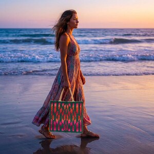 May include: A woman walking on a beach at sunset, carrying a green tote bag with a red and pink abstract design. She wears a long, flowing dress with a floral pattern. The ocean waves and sky are visible in the background.