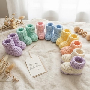 Puede incluir: Una colección de botines de bebé de ganchillo de colores pastel, dispuestos en semicírculo. Los botines son de diferentes colores: lila, verde, rosa, azul, amarillo y melocotón. Una etiqueta dice: "Welcome Little One!" con "xo".