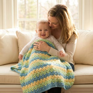 May include: A baby wrapped in a handmade crochet blanket, held by a person on a beige sofa. The blanket features horizontal stripes of blue, yellow, and green. The baby is smiling. The person is wearing a long-sleeved cream-colored shirt.