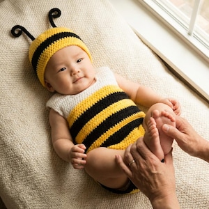 May include: A baby dressed in a handmade crochet bee outfit, featuring a hat with antennae and a yellow and black striped bodysuit. The baby is lying on a beige surface, with hands gently touching the baby's feet.