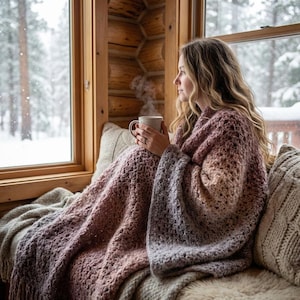 Op de afbeelding: Een persoon gehuld in een grote, gefranjerde, ombre gehaakte deken, die overgaat van roze naar grijs. Ze zitten bij een raam en houden een mok vast, met uitzicht op een besneeuwde omgeving. De deken is een warm, getextureerd item.