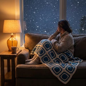 Puede incluir: Una persona sentada en un sofá marrón, envuelta en una manta de ganchillo azul y crema, mirando por una ventana la nieve que cae. Una lámpara de cristal ámbar y una taza están sobre una mesa auxiliar de madera.