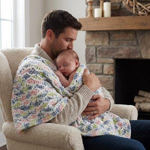 May include: A baby wrapped in a pastel-coloured crocheted blanket, held by a person in a beige jumper. The blanket features a pattern of blue, pink, green, and white. The person is seated in a cream-coloured armchair, with a stone fireplace in the background.