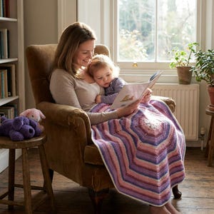 May include: A woman and child are seated in a brown armchair, reading a book together. A colorful, geometric-patterned blanket covers their laps. A small wooden stool holds two plush toys. The room has a window and a bookshelf.