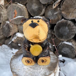 May include: A hand-carved wooden bear figurine with a light brown face and dark brown accents. The bear is holding a gold-colored, glittery ornament. The figurine is sitting on a tree stump in a snowy environment.