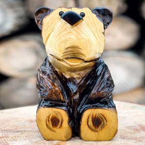 May include: A wooden bear carving with a brown and black finish. The bear is sitting with its paws in front of it. The carving is detailed with a carved face and eyes.