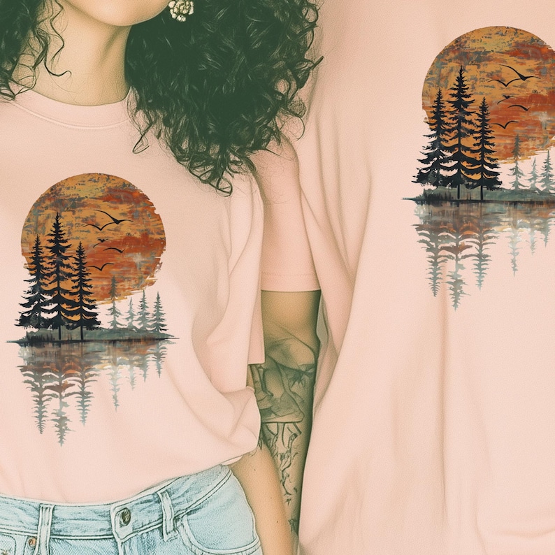 May include: Light pink crew neck t-shirts featuring a nature-themed graphic. The design includes a sunset, pine trees, and birds reflected in water. The shirts are made of a soft material.