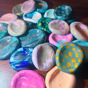 May include: A collection of small, round, concave clay discs featuring various finishes including marbled swirls, solid bright colors, and gold-painted checkered patterns. Each disc has a smooth, glossy surface with unique color blends.