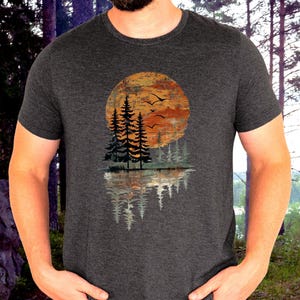 May include: Dark gray t-shirt featuring a nature-themed graphic. The design includes a sunset with trees and birds, reflected in water. The graphic is in shades of orange, brown, and gray.