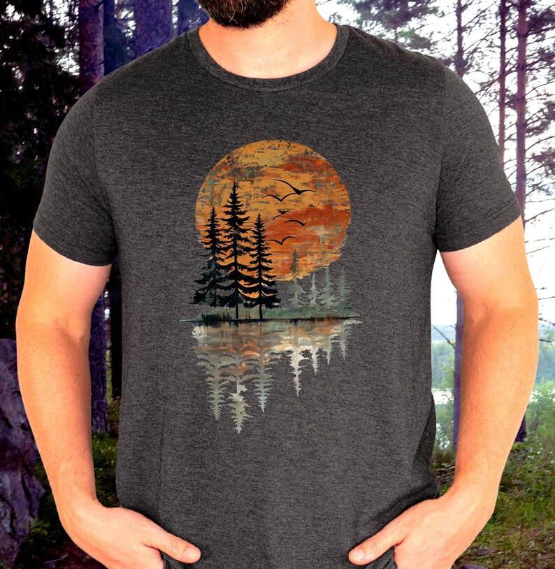 May include: Dark gray t-shirt featuring a nature-themed graphic. The design includes a sunset with trees and birds, reflected in water. The graphic is in shades of orange, brown, and gray.