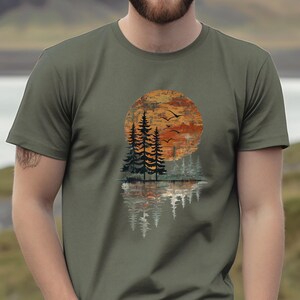 May include: Olive green t-shirt featuring a sunset scene with trees, birds, and a water reflection. The design includes a large orange and brown sun, silhouetted trees, and flying birds, creating a nature-inspired graphic.
