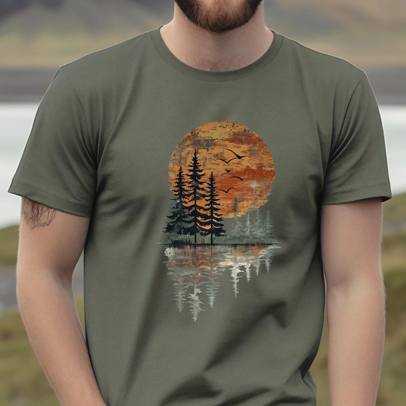 May include: Olive green t-shirt featuring a sunset scene with trees, birds, and a water reflection. The design includes a large orange and brown sun, silhouetted trees, and flying birds, creating a nature-inspired graphic.