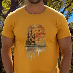May include: Mustard yellow t-shirt featuring a nature-themed graphic. The design includes a large orange and brown sun, silhouetted pine trees, and birds in flight. The trees are reflected in a body of water below.