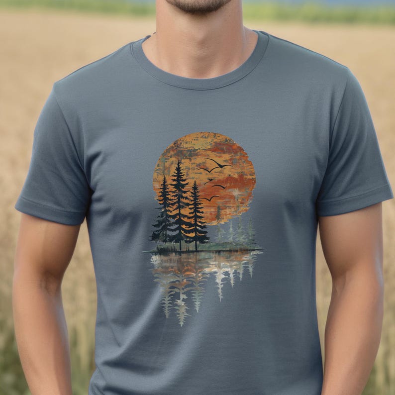 May include: A slate blue t-shirt featuring a nature-themed graphic. The design includes a large orange and yellow sun, silhouetted pine trees, and flying birds, with a mirrored reflection below. The shirt is made of a soft material.