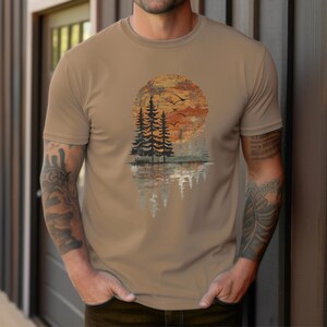 May include: A tan-colored t-shirt featuring a nature-themed graphic. The design includes a sunset, pine trees, birds, and their reflection in water. The artwork is in shades of orange, brown, and gray.