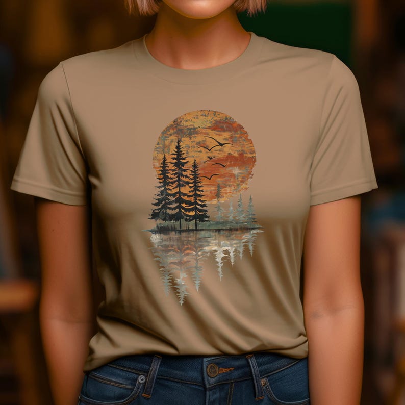 May include: Tan short-sleeve t-shirt featuring a nature-themed graphic. The design includes a sunset, trees, birds, and their reflection in water. The artwork is in shades of orange, brown, and green.