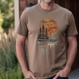 May include: A tan-colored t-shirt featuring a sunset scene with pine trees and birds in flight. The design includes a large orange sun, silhouetted trees, and their reflection in the water below. The shirt is a short-sleeved crew neck.