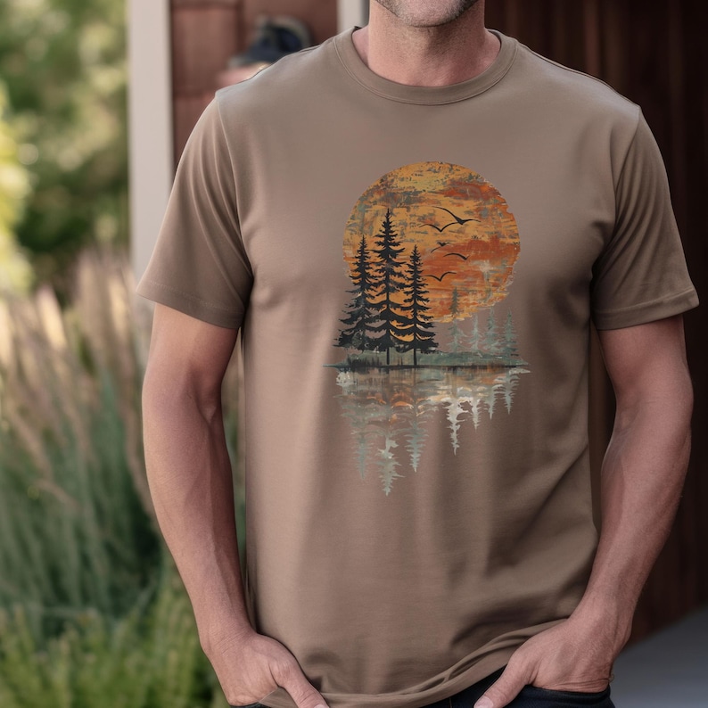 May include: A tan-colored t-shirt featuring a sunset scene with pine trees and birds in flight. The design includes a large orange sun, silhouetted trees, and their reflection in the water below. The shirt is a short-sleeved crew neck.