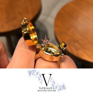Puede incluir: Un anillo dorado con una tapa con bisagras, que revela un pequeño dije en forma de estrella. El anillo tiene un diseño detallado y está abierto, mostrando el interior. El anillo es parte de la colección "Vei Jewelry".