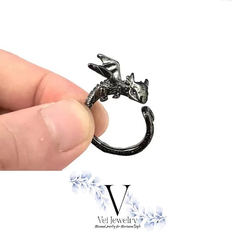 Puede incluir: Un anillo de drag&oacute;n gris oscuro. El drag&oacute;n tiene alas y escamas detalladas. El anillo es abierto, lo que permite ajustar la talla. El anillo est&aacute; sujeto por los dedos de una persona. Es una joya.