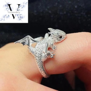 Puede incluir: Un anillo de plata con forma de drag&oacute;n con detalles intrincados, incluyendo alas y escamas texturizadas. El anillo est&aacute; envuelto alrededor de un dedo, mostrando la joyer&iacute;a de tem&aacute;tica fant&aacute;stica. El anillo es de Vel Jewelry.