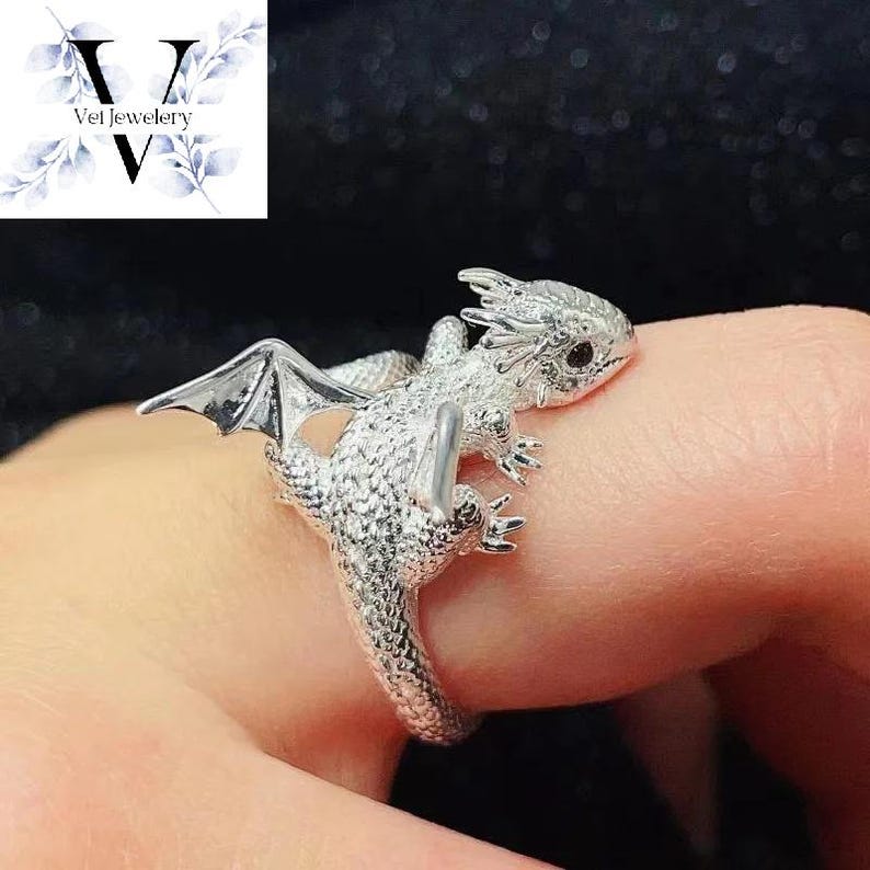 Puede incluir: Un anillo de plata con forma de drag&oacute;n con detalles intrincados, incluyendo alas y escamas texturizadas. El anillo est&aacute; envuelto alrededor de un dedo, mostrando la joyer&iacute;a de tem&aacute;tica fant&aacute;stica. El anillo es de Vel Jewelry.