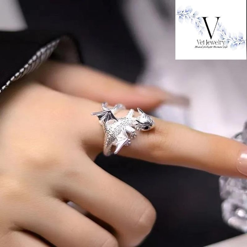 Puede incluir: Anillo de plata con forma de drag&oacute;n, con detalles intrincados, que presenta un drag&oacute;n con alas extendidas y un cuerpo texturizado. La cabeza del drag&oacute;n est&aacute; colocada en el dedo, con un ojo negro. Una joya.