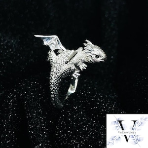 Puede incluir: Un anillo de drag&oacute;n plateado con escamas detalladas, alas y una cabeza peque&ntilde;a. El drag&oacute;n est&aacute; posado en una banda de anillo. El fondo es negro. Es una joya.