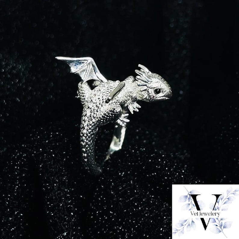 Puede incluir: Un anillo de drag&oacute;n plateado con escamas detalladas, alas y una cabeza peque&ntilde;a. El drag&oacute;n est&aacute; posado en una banda de anillo. El fondo es negro. Es una joya.