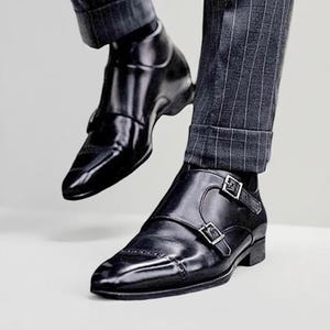 May include: Black leather double monk strap shoes with silver buckles. The shoes have a pointed toe and are worn with gray pinstripe trousers. The shiny shoes are a classic style.