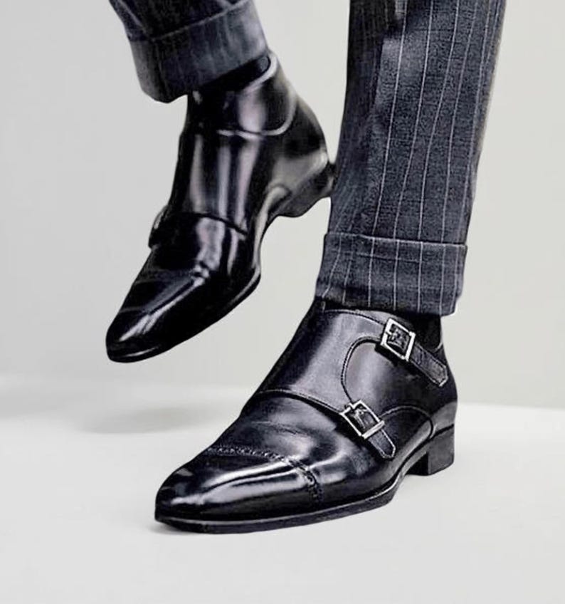 May include: Black leather double monk strap shoes with silver buckles. The shoes have a pointed toe and are worn with gray pinstripe trousers. The shiny shoes are a classic style.