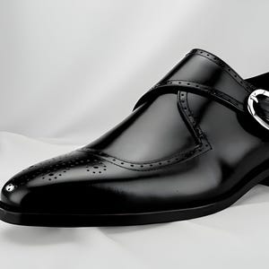 May include: A single black leather shoe with a glossy finish. The shoe has a decorative strap with a silver buckle and detailed perforations on the toe. The shoe is set against a white backdrop.
