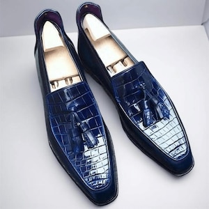 May include: A pair of navy blue leather loafers with a crocodile-textured vamp and a tassel detail. The shoes have a classic slip-on design and are displayed on a white surface. The shoes have wooden shoe trees inside.