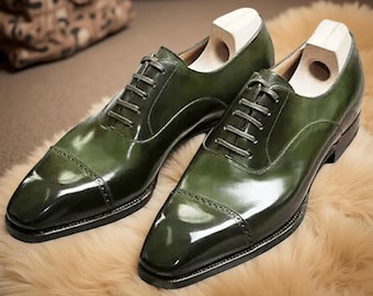 May include: A pair of green leather dress shoes with a glossy finish. The shoes have a classic Oxford design with a cap toe and lace-up closure. Wooden shoe trees are inserted inside the shoes. The shoes are placed on a light brown, textured surface.