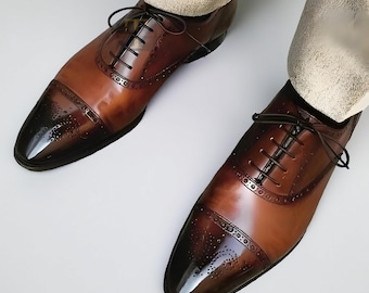 May include: A pair of brown leather dress shoes with a two-tone design. The toe caps are a darker shade of brown, and the shoes feature intricate perforations and black laces. The shoes are worn with beige pants.